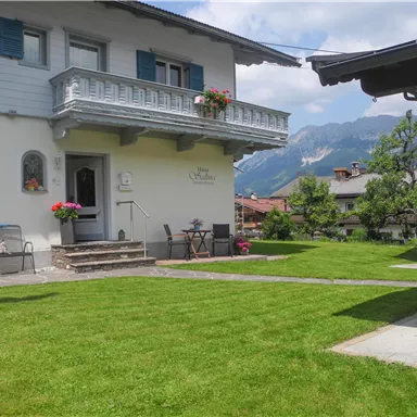 Ein schönes Haus mit einem gepflegten Garten und blühenden Blumen. Im Hintergrund sind Berge und ein klarer Himmel zu sehen.