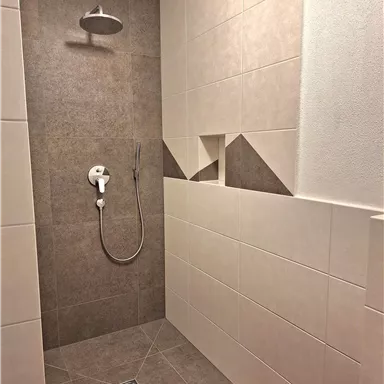 Eine moderne Dusche mit hellen Fliesen und einem grauen Akzent. Die Duschinstallationen sind minimalistisch und elegant gestaltet.