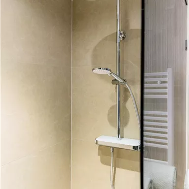 A modern shower with a large rain shower head and a clear, elegant design. In the background, bright tiles and a towel holder are visible.