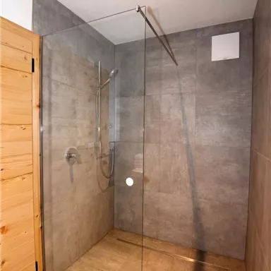 Eine moderne Duschkabine mit gläserner Trennwand und grau gefliesten Wänden. Der Boden ist aus hellem Material und die Holzdetails verleihen einen warmen Touch.