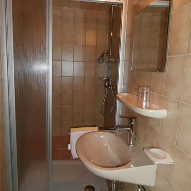 Ein Badezimmer mit einer Dusche und einem kleinen Waschbecken. Die Wände sind mit beige Fliesen gefliest.