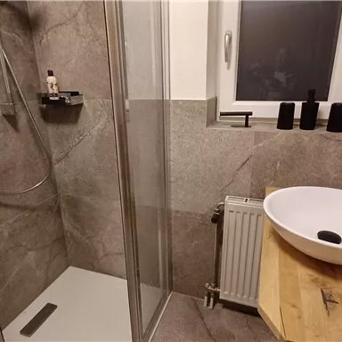 Ein modernes Badezimmer mit einer Glasdusche und einem Waschbecken auf einem Holzregal. Die Wände sind in sanften Grautönen gehalten.