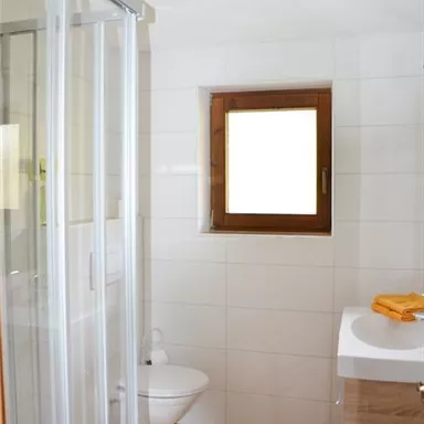 Ein modernes Badezimmer mit einer Dusche und einemWC. Der Raum hat helle Fliesen und ein Fenster mit Holzrahmen.