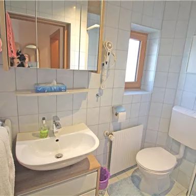 Ein helles Badezimmer mit einem Waschtisch, Waschbecken und einer Dusche. Die Wände sind gefliest und es gibt ein Fenster, das natürliches Licht hereinlässt.