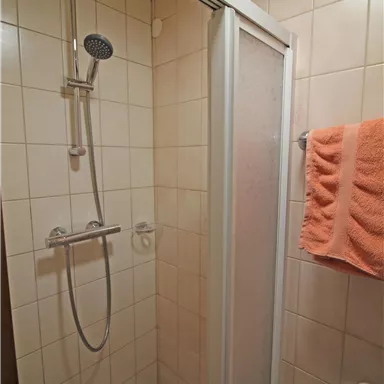 Eine einfache Dusche in einem Badezimmer mit weißen Fliesen. Ein Handtuch in Orange hängt neben der Dusche.