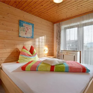 Ein gemütliches Schlafzimmer mit Holzvertäfelung und einem großen Bett. Bunte Kissen und ein Fenster mit Vorhängen sorgen für eine einladende Atmosphäre.