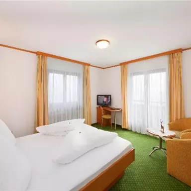 A bright hotel room with a double bed and a cozy armchair. There are large windows that let in plenty of light and a television.