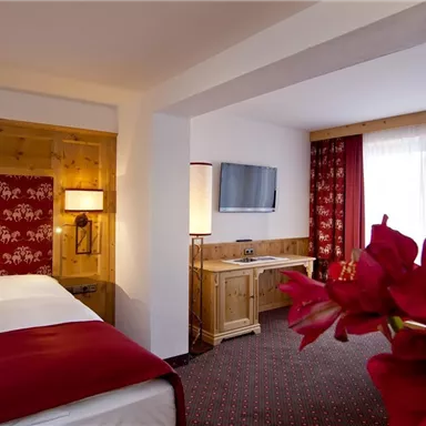 A cozy hotel room with wooden paneling and red accents. The room features a bed, a television, and a window with curtains.