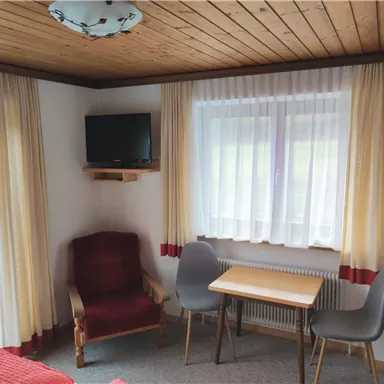 Ein gemütliches Zimmer mit einem roten Bett, einem kleinen Tisch und zwei Stühlen. An der Wand hängt ein Fernseher und die Fenster sind mit hellen Vorhängen dekoriert.