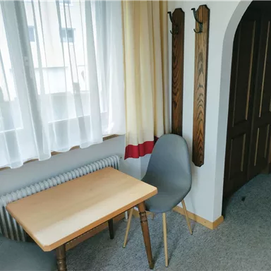 Ein kleines Zimmer mit einem Tisch und einem Stuhl. Der Raum hat große Fenster und eine gemütliche Atmosphäre.