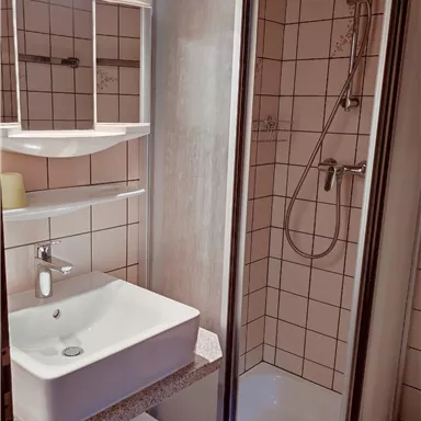 Ein Badezimmer mit einer modernen Dusche und einem Waschbecken auf einem minimalistischen Regal. Die Fliesen sind hell und die Beleuchtung ist freundlich.