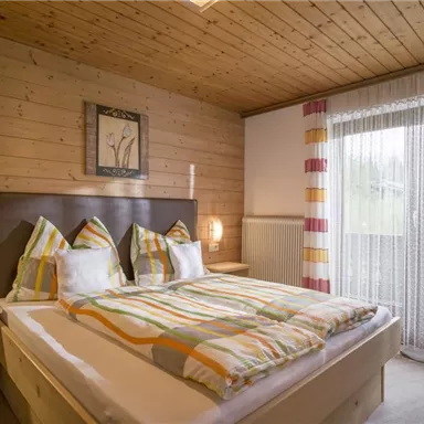 Ein gemütliches Schlafzimmer mit Holzvertäfelung und einem großen Bett. Es gibt einen Balkon mit hellen Vorhängen und dekorativen Kissen.