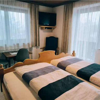 Ein gemütliches Schlafzimmer mit zwei Betten und großen Fenstern. Es gibt einen Tisch, Stühle und einen Fernseher im Hintergrund.