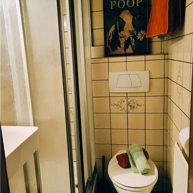 Ein kleines Badezimmer mit einer Dusche und einer Toilette. An der Wand hängt ein humorvolles Schild.