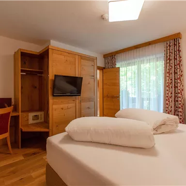 Ein gemütliches Hotelzimmer mit einem Doppelbett, einem Schreibtisch und einem Fernseher. Große Fenster sorgen für viel Tageslicht.