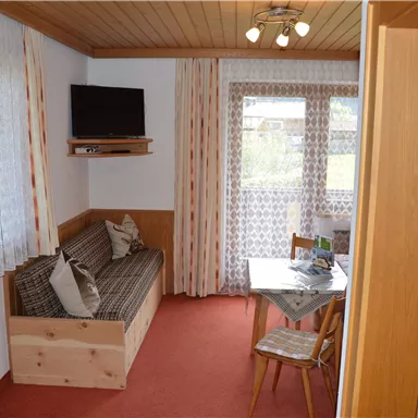 Ein gemütlicher Wohnraum mit einem Sofa, einem Tisch und einem Fernseher. Große Fenster sorgen für viel Licht und Blick nach draußen.