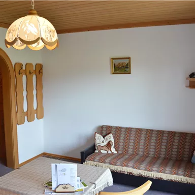 Ein gemütliches Wohnzimmer mit einem Sofa, einem Tisch und einem Fernseher auf einem Regal. Die Wände sind hell, und eine schöne Lampe hängt von der Decke.