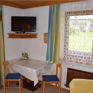 Ein gemütlicher Wohnraum mit einem Tisch und zwei Stühlen. An der Wand hängt ein Fernseher und ein Fenster lässt viel Tageslicht herein.