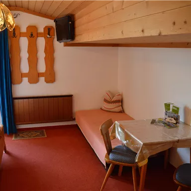 Ein gemütliches Zimmer mit Holzdetails und einem bequemen Einzelbett. Am Tisch stehen zwei Stühle und ein Informationsheft.