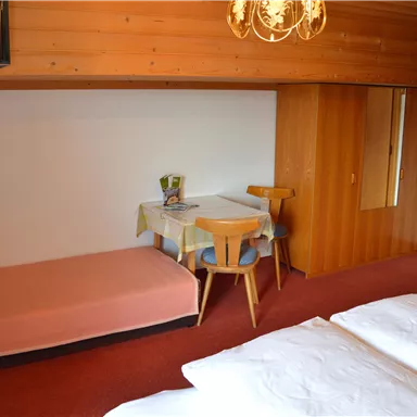 Ein gemütliches Zimmer mit Holzwänden, einem Doppelbett und einem kleinen Esstisch. Im Hintergrund befindet sich ein zusätzliches Schlafsofa.