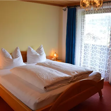 Ein gemütliches Schlafzimmer mit einem Doppelbett und zwei großen Kissen. Das Zimmer hat eine helle Atmosphäre und ein Fenster mit Vorhängen.