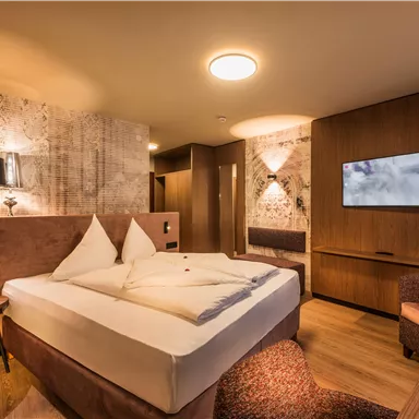 A modern hotel room with a large bed, elegant wood paneling, and a wall-mounted TV. Cozy furniture and warm lighting create an inviting atmosphere.