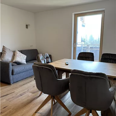 Ein modernes Wohnzimmer mit einem grauen Sofa und einem Holztisch. Große Fenster bieten viel Tageslicht und Zugang zu einem Balkon.