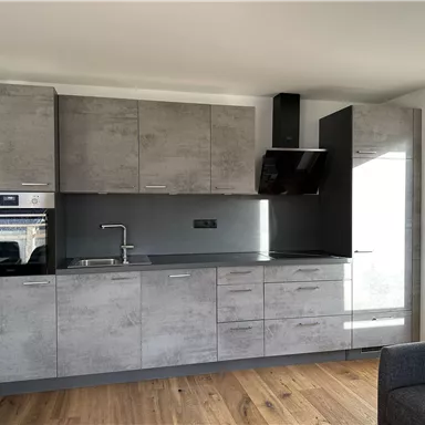 Eine moderne Küche mit grauen Schränken und schwarzen Geräten. Der Boden ist aus Holz und es gibt große Fenster für viel Licht.