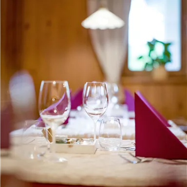 Ein schön gedeckter Tisch mit Gläsern und lila Servietten. Die warme Holzdecke und das Fenster schaffen eine gemütliche Atmosphäre.