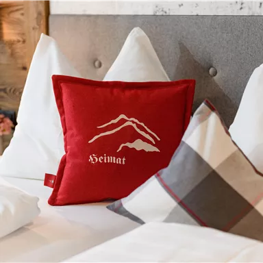 A cozy bed with decorative cushions. The red cushion features the inscription "Home" and has a mountain motif.