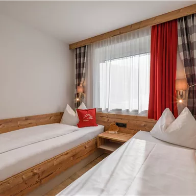 A cozy room with two single beds and wooden details. Bright curtains and red accents create an inviting atmosphere.