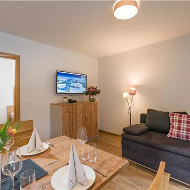A cozy living room with modern furniture. A dining table is placed next to a couch with cushions and a television.