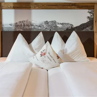 A cozy bedroom with a large bed and decorative pillows. In the background, there is a picture of mountains.