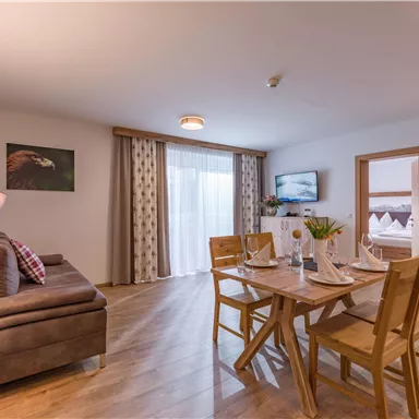 A modern living room with a cozy sofa and a wooden dining table. Large windows let in plenty of daylight and provide a view outside.