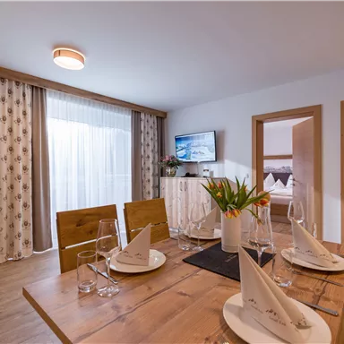 A modern dining room with a wooden table, set with plates and glasses. Large windows let in plenty of light and a television is mounted on the wall.