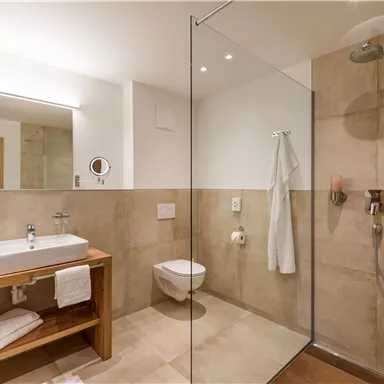 A modern bathroom with a glass shower and light, neutral color tone. The sink is on a wooden shelf with fresh towels.