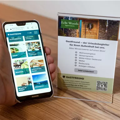 A smartphone displays the website of the Gastfreund app, which provides information about culinary offerings and leisure activities. Next to it stands an information sign for the apartment complex Echt Woods.