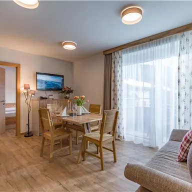A cozy living room with a wooden table and chairs. Large windows provide plenty of daylight and a pleasant atmosphere.