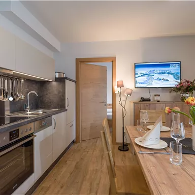A modern kitchen with a dining area and a large table. The room is bright and inviting, with a television on the wall.