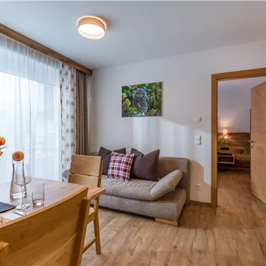 A cozy living space with a sofa, a table, and a window that lets in plenty of daylight. On the wall hangs a picture of an owl, and there are fresh flowers on the table.