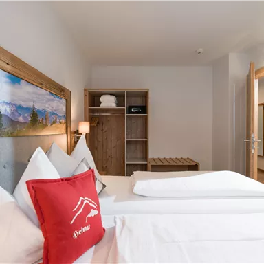 A cozy hotel room with a double bed and decorative cushions. In the background, a wooden shelf and a wardrobe are visible.