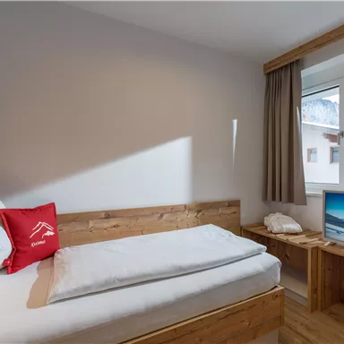 A cozy room with a single bed, wooden furniture, and a TV. Natural light comes through the window and offers a view outside.