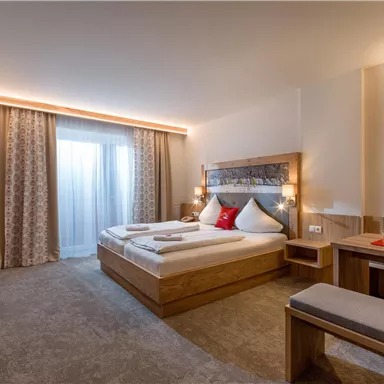 Ein modernes Hotelzimmer mit einem Doppelbett und einer gemütlichen Sitzecke. Große Fenster lassen viel Tageslicht herein und die Einrichtung ist aus hellem Holz.