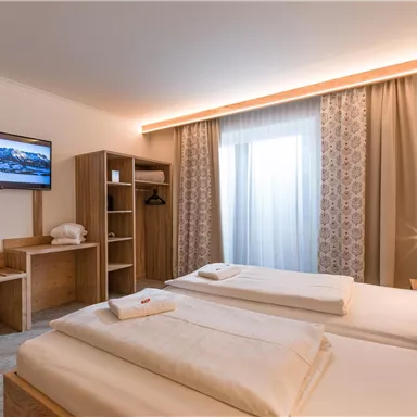 Ein modernes Hotelzimmer mit zwei Betten und einem Fernseher. Große Fenster lassen viel Tageslicht herein und sorgen für eine helle Atmosphäre.