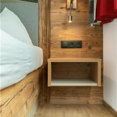 A cozy bedroom with a wooden bed and a bedside lamp.  
On the wall, there are power outlets and a small table.