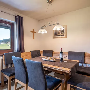 Ein einladender Essbereich mit einem Holztisch und sechs Stühlen. Große Fenster bieten Blick auf die Landschaft, und eine Flasche Wein steht auf dem Tisch.