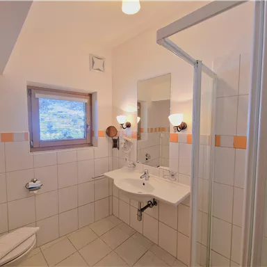 Ein helles Badezimmer mit Dusche, Waschbecken und Spiegel. Es hat weiße Fliesen und ein Fenster, das Licht hereinlässt.