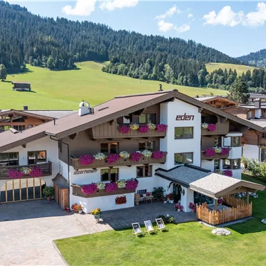 Ein gemütliches Hotel mit blühenden Balkonkästen inmitten einer grünen Landschaft. Im Hintergrund sind sanfte Hügel und ein klarer Himmel sichtbar.