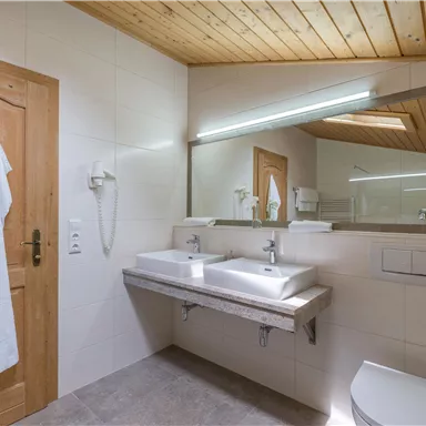 Ein modernes Badezimmer mit zwei Waschbecken und einem großen Spiegel. Die Wände sind hell und die Decke aus Holz.