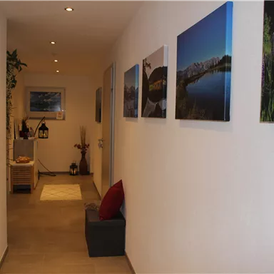 Ein schmaler Flur mit weißen Wänden und mehreren Bildern an der Wand. Am Ende des Flurs gibt es einen kleinen Tisch und eine Pflanze.